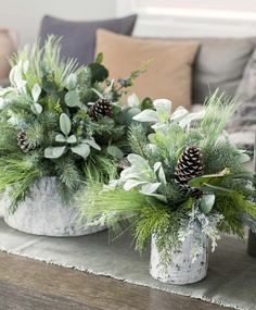 two white vases filled with greenery and pine cones on a table in front of a couch