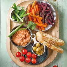 an assortment of vegetables and dips on a wooden platter with bread, tomatoes, carrots, celery, olives