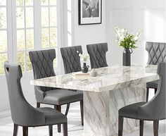 a marble dining table surrounded by grey chairs and vases with flowers on it in a white room