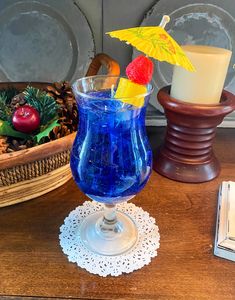a blue drink in a glass with an umbrella and pineapple on the top sits next to a candle