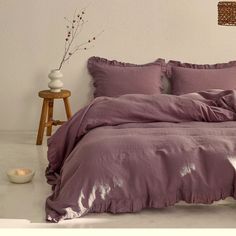 a bed with purple sheets and pillows in a white room next to a table lamp