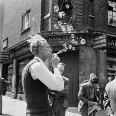 an old man blowing bubbles on the street while others walk by in front of him