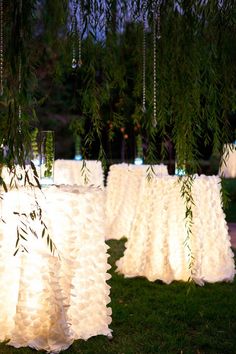 several tables covered in white cloths under trees with lights hanging from the branches above them
