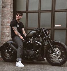 a man sitting on top of a black motorcycle next to a brick building and wearing sunglasses