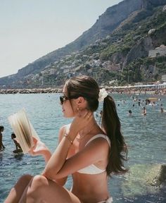 a woman sitting on the edge of a body of water with a book in her hand