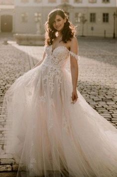a woman in a wedding dress standing on a cobblestone road with her arms behind her back