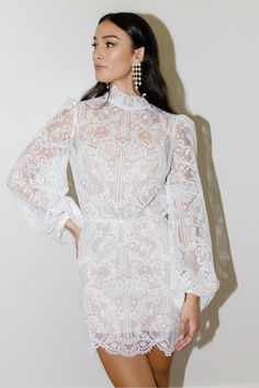 a woman wearing a white lace dress with long sleeves and an open back, standing in front of a wall