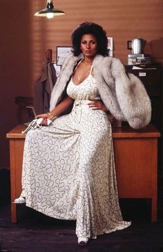 a woman in a white dress and fur coat sitting on a desk with her hands on her hips