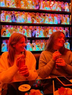 two women sitting at a table with drinks in front of colorful bottles on the wall