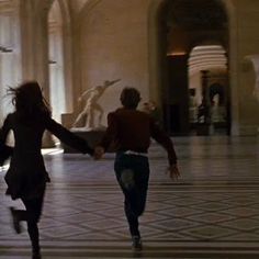 two people are running in the middle of a room with statues on either side of them