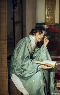 a woman sitting on the floor reading a book while wearing a green kimono and holding an open book in her hand