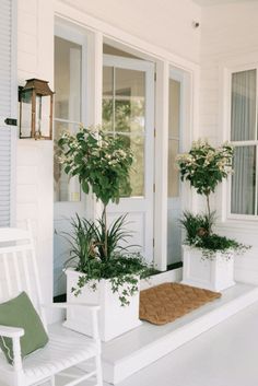 two white chairs sitting on the front porch with plants in planters and a door mat