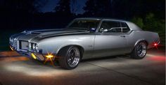 an old muscle car is parked in the driveway at night with its headlights turned on