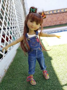 a doll with long brown hair and blue overalls standing next to a white fence