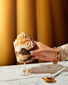 a hand holding a glass filled with dessert on top of a marble table next to gold spoons