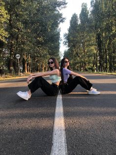 two young women sitting on the side of an empty road with trees in the background