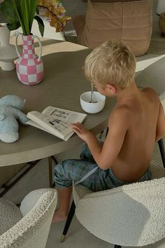 a young boy sitting at a table reading a book and drinking from a coffee cup