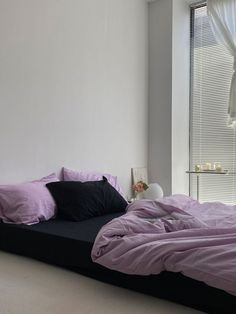 a bed with purple sheets and pillows in a white room next to a large window