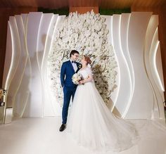 a bride and groom standing in front of a floral wall at their wedding reception venue