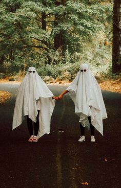 two people in white ghost costumes holding hands on a road with trees and leaves behind them