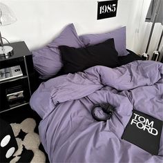 an unmade bed with purple sheets and black pillows in a white room next to a lamp