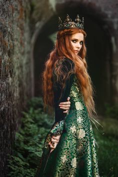 a woman with long red hair wearing a green dress and crown standing in the woods