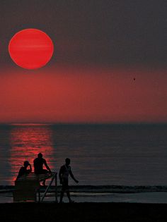 three people sitting on a bench at the beach watching the sun go down over the ocean