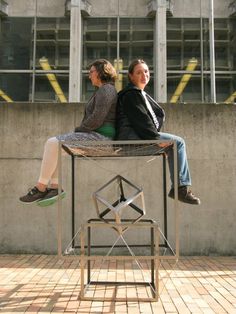two women sitting on top of a metal table next to each other in front of a building