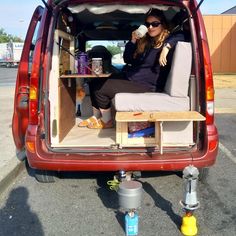 a woman sitting in the back of a red van drinking from a cup and holding a coffee mug