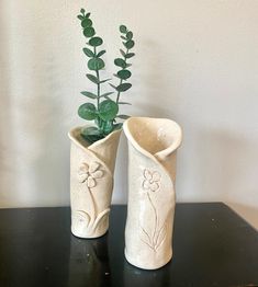 two white vases with plants in them on a black table next to a wall