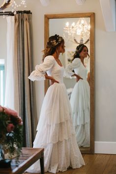 a woman standing in front of a mirror wearing a white wedding dress with ruffles