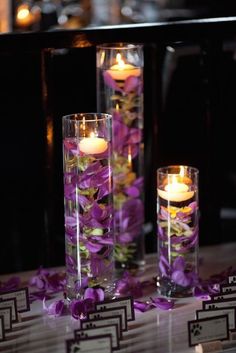 three tall clear vases filled with purple flowers and lit candles on a white table