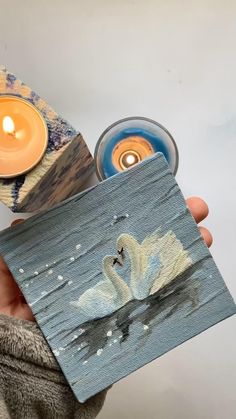 a person holding up a small piece of art with a candle in it and a swan painting on the front