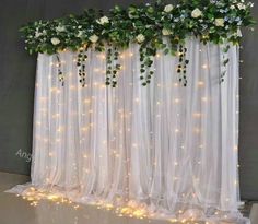 a white curtain with flowers and greenery hanging from it's sides is lit up by fairy lights