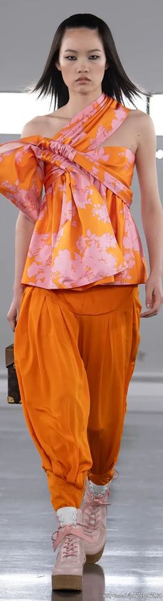 a woman walking down a runway wearing an orange top and matching pants with pink shoes