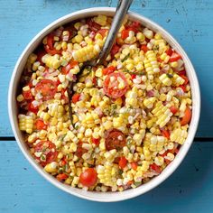 a bowl filled with corn and tomatoes on top of a blue wooden table next to a spoon
