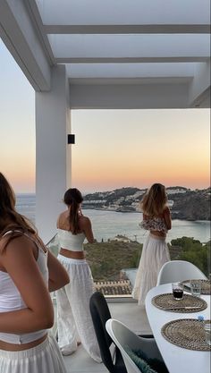 three women in white dresses standing on a balcony looking at the water and hills behind them