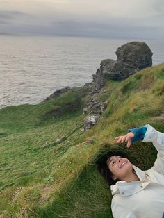 a woman laying on top of a lush green hillside next to the ocean with her arms outstretched