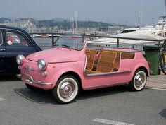 an old pink car is parked next to a boat in the water with its doors open