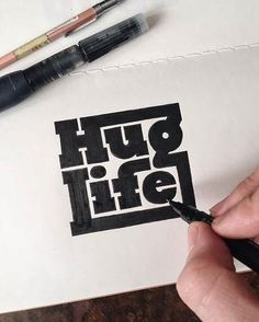 someone is drawing on paper with black marker and some pens in front of them, while another hand holds a pen