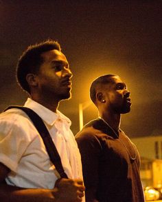 two men standing next to each other in the street at night with one looking off into the distance