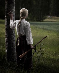 a woman standing next to a tree in a field holding a stick and wearing a white shirt