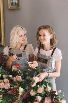 two women standing next to each other with flowers in front of them and holding scissors