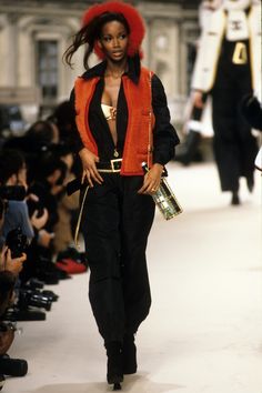 a woman walking down a runway with an orange hat on her head and black pants