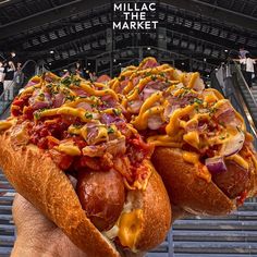 two hot dogs covered in cheese and toppings are held up by someone's hand