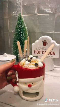 a person holding a cup with hot chocolate and marshmallows in front of a christmas tree