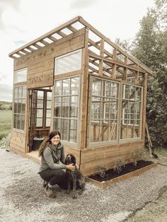 a woman kneeling down next to a dog in front of a small wooden structure with windows