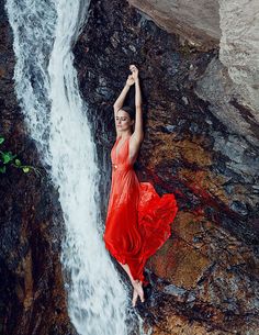 a woman in a red dress standing on rocks next to a waterfall and holding her arms above her head
