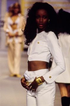 a model walks down the runway wearing white clothes and high heels with gold accessories on her hands