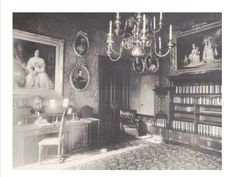 an old black and white photo of a room with pictures on the wall, chandelier, bookshelves, and desk
